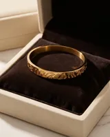 Esclava rígida de acero con baño de oro, con diseño grabado en relieve. Se presenta junto a una pulsera de cadena fina de eslabones.