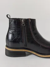 Bota corta de cuero negro con textura de reptil, diseño tipo chelsea con elásticos laterales y costuras a la vista en tono contrastante. Cuenta con suela plana de goma con detalle de franja en color natural.