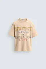 Camiseta relaxed fit de punto color beige con efecto lavado, cuello redondo y manga corta. Cuenta con estampado frontal con texto en tonos amarillo, azul y rojo.