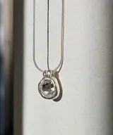 Collar plateado con dije circular con piedra transparente facetada. Cadena fina de eslabones.