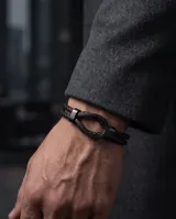 Pulsera tipo esclava compuesta por una doble tira de cuero trenzado negro y un cierre central de acero en forma de herradura.
