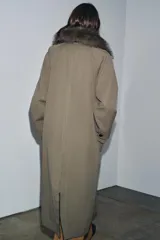 Trench reversible de corte recto con cuello de solapa y aplicación de pelo sintético extraíble. Presenta cierre frontal con botones, manga larga y bolsillos laterales ocultos en las costuras.