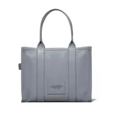 Cartera tipo tote de cuero granulado en color gris, con diseño estructurado y asas superiores. Presenta el logo estampado en el frente con la inscripción The Tote Bag Marc Jacobs.