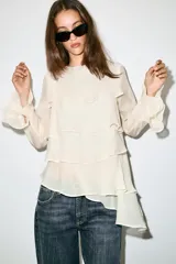 Blusa marrón de cuello redondo y manga larga, con volantes horizontales superpuestos y bajo asimétrico. Cuenta con detalle de abertura y botón en la espalda.