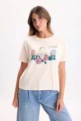 Remera manga corta de color crudo, con cuello redondo y estampa frontal ilustrada de Alicia en el País de las Maravillas rodeada de flores.