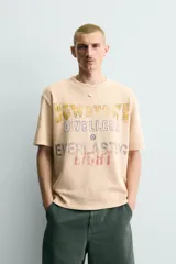 Camiseta relaxed fit de punto color beige con efecto lavado, cuello redondo y manga corta. Cuenta con estampado frontal con texto en tonos amarillo, azul y rojo.