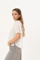 Blusa blanca de manga corta con cierre frontal de botones y detalle de pliegues verticales en la parte delantera. Presenta un corte recto y relajado, confeccionada en una tela liviana con textura.