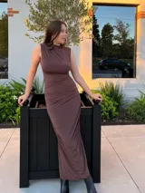 Vestido largo de color marrón, con diseño sin mangas y cuello alto. Presenta un detalle drapeado en el lateral que recorre la silueta, aportando textura y un ajuste entallado.