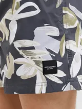 Short de baño con estampado floral abstracto en tonos verdes, beige y blancos sobre base gris oscuro. Presenta cintura elástica con cordón ajustable y bolsillos laterales.