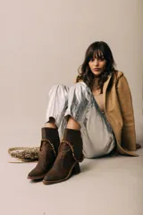 Botas de cuero marrón con punta cuadrada levemente elevada y taco ancho. Presentan una caña corta con detalle de costuras expuestas y una tira trenzada decorativa alrededor del tobillo.