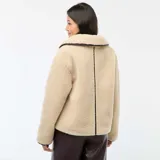 Campera de efecto borreguito en color beige, con cuello amplio y cierre frontal de botones negros. Presenta ribetes negros en contraste en el cuello, la apertura frontal y los puños. Marca Kiabi.