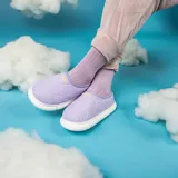 Pantuflas acolchadas de diseño tipo slip-on en color lila, con costuras onduladas y suela de goma blanca. Presentan el logo de la marca bordado en amarillo en la parte superior.