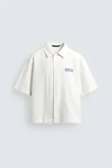 Camisa de corte relajado confeccionada en tejido de algodón con efecto lavado. Presenta cuello solapa, manga corta y cierre frontal con botones. Incluye un estampado a contraste en el frente.