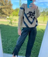 Sweater tostado de lana con cuello redondo y mangas largas. Presenta apliques de cuero negro en forma de V en el frente y bordado de una rosa negra en el pecho. El pantalón es de cuero ovino negro con corte slouchy y aberturas en el ruedo.