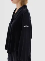 Camisa de terciopelo color negro, con cuello clásico, cierre frontal oculto y mangas largas con puños elásticos. Presenta un diseño holgado con cordón ajustable en el ruedo.