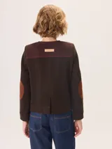 Chaqueta de paño color marrón oscuro, de corte clásico y cuello redondo, con cierre frontal dorado. Presenta bolsillos rectangulares con ribetes en color rojizo y parches de pana a rayas en los hombros.
