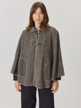 Capa de tejido de punto en color gris melange, con diseño abierto y cierre frontal mediante un botón tipo duffle de madera con detalle en cuero. Presenta bolsillos laterales amplios y bordes con costuras a la vista.