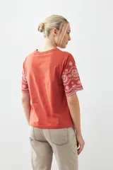 Remera de algodón color terracota con detalles de bordado broderie blanco en mangas y ruedo. Presenta cuello redondo y mangas cortas.