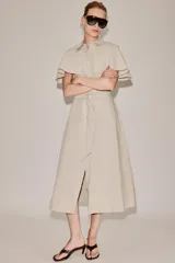 Vestido camisero midi de algodón con diseño de capa, cuello solapa, manga corta tipo capa y trabillas en los hombros. Presenta cierre frontal con botones, bolsillos delanteros y cinturón ajustable del mismo tejido con lazada. El bajo tiene un acabado en línea evasé.