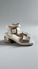Sandalias de cuero color blanco y dorado, con taco ancho intervenido con arte pintado a mano, tira frontal metalizada, tira al tobillo con hebilla metálica y plataforma baja.