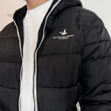 Campera inflada negra con capucha, cierre frontal blanco y puños elásticos acanalados. Presenta el logo de la marca Santa Barbara Outfitters bordado en el pecho.