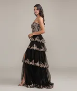 Vestido largo de fiesta con escote corazón y corset estructurado. Presenta un diseño de capas de tul con volados y bordados brillantes en tonos dorados sobre base negra. Incluye un tajo lateral profundo en la falda.