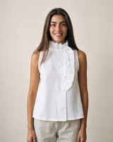 Blusa blanca de lino sin mangas, con cuello alto plisado, cierre lateral asimétrico con botones forrados al tono y volados decorativos.
