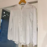 Camisa blanca de manga larga con cuello mao y cierre frontal de botones. Presenta detalles de pliegues fruncidos en el canesú delantero y los hombros.