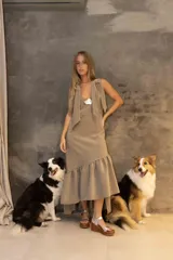 Vestido midi color arena con breteles anchos que se atan en los hombros, escote en V y falda con volado asimétrico en el ruedo. Incluye un aplique decorativo metálico en el centro del pecho.