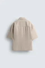 Camisa de corte relajado color beige, con cuello bowling, manga corta y cierre frontal con botonadura oculta por solapa. Presenta aberturas laterales en el bajo.