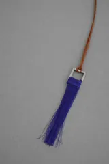 Collar largo con cordón de cuero marrón y dije colgante compuesto por una pieza metálica cuadrada y una borla de hilos azul eléctrico.
