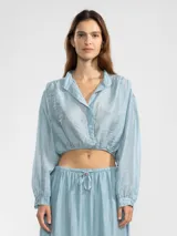 Blusa corta de tejido arrugado liviano, con cuello camisero, cierre frontal de botones y mangas largas abullonadas con puños. Presenta un ruedo elastizado que le otorga una silueta amplia y relajada.