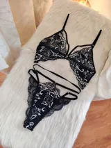 Conjunto de lencería compuesto por un bralette triangular y una tanga, confeccionados en encaje negro con detalles florales en color hueso. El bralette presenta breteles ajustables y diseño con tiras decorativas, mientras que la tanga cuenta con doble tira lateral regulable.
