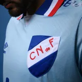 Camiseta de fútbol celeste del Club Nacional de Football, marca Umbro, con cuello en V y detalles en blanco y rojo. Incluye escudo del club, logo de Umbro y sponsors.