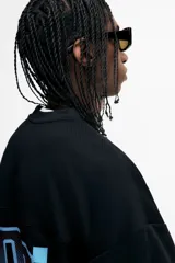 Remera de malla negra con cuello en V, mangas cortas y hombros caídos. Presenta un estampado frontal con la frase "THUNDER X RACING TEAM" y un diseño gráfico en tonos rosa, azul y celeste con la palabra "RACING" y un patrón de cuadros.