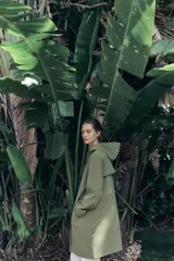 Chubasquero verde militar de cuello subido con capucha ajustable con cordones. Tiene manga larga, bolsillos delanteros con solapa y botón a presión oculto, y cierre frontal con cremallera oculta y botones a presión.