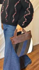 Bolso tipo matera confeccionado en cuero vacuno, con diseño estructurado y manija superior integrada. Presenta un cuerpo con franjas diagonales en gamuza de colores contrastantes (amarillo, verde y azul) sobre una base de cuero marrón, con detalles de costuras artesanales en los bordes.