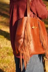 Bolso tipo tote de cuero color marrón, con diseño de flecos laterales y cierre frontal mediante un botón de guampa. Cuenta con doble asa de hombro.