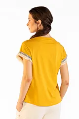 Remera de manga corta color mostaza, confeccionada en 100% algodón, con cuello redondo y detalles de flecos y guardas geométricas en las mangas.