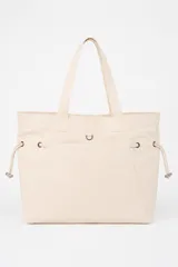 Bolso tipo tote bag color beige con asas superiores de hombro, cordones ajustables a los lados con detalles metálicos y un pequeño anillo frontal.