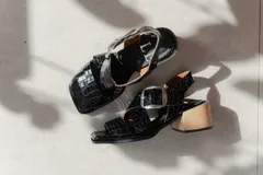 Sandalias de cuero negro con textura de reptil, diseño de punta cuadrada y taco bloque de madera. Presentan tiras cruzadas en el empeine y una hebilla metálica grande decorativa.