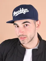Gorro tipo snapback de color azul marino con visera plana y bordado frontal en blanco con la palabra "Brooklyn" en estilo cursiva.