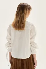 Camisa blanca de corte recto, con cuello clásico y mangas largas abullonadas que presentan detalles en punto smock en los puños. Incluye un pequeño bordado en el pecho.