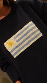 Sweater de hilo de algodón color verde militar, con cuello redondo y manga larga. Presenta un diseño tejido de la bandera uruguaya en el frente.