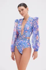 Body de manga larga con estampado floral en tonos azules y rosados. Presenta un escote profundo en V, detalle de nudo en el centro del pecho y en la cintura, aberturas laterales y mangas largas transparentes con hombros estructurados y puños elásticos.