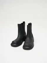 Bota tipo Chelsea de caña media, confeccionada en cuero vacuno negro con acabado mate. Presenta paneles laterales elásticos, tirador trasero y una suela robusta con plataforma delantera y taco cuadrado.