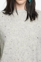 Sweater de punto color gris melange, confeccionado en una mezcla de lana y alpaca. Presenta un diseño de corte recto con escote redondo, mangas largas tipo murciélago con puños anchos y detalles de piedras brillantes aplicadas en toda la prenda.