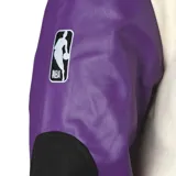Campera bomber de algodón color crema con mangas de cuero sintético color violeta. Tiene el logo de Los Angeles Lakers bordado en el pecho y parches en los codos.