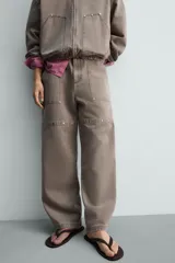 Pantalón de corte relajado en color marrón con efecto lavado. Presenta bolsillos frontales tipo cargo, cierre con cremallera y botón, y una línea horizontal de tachas metálicas a tono en la parte superior de las piernas.