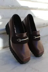 Mocasín de cuero vacuno color bordó con suela track negra de goma. Presenta un diseño de punta cuadrada con un herraje metálico plateado sobre una banda de gamuza en la capellada.